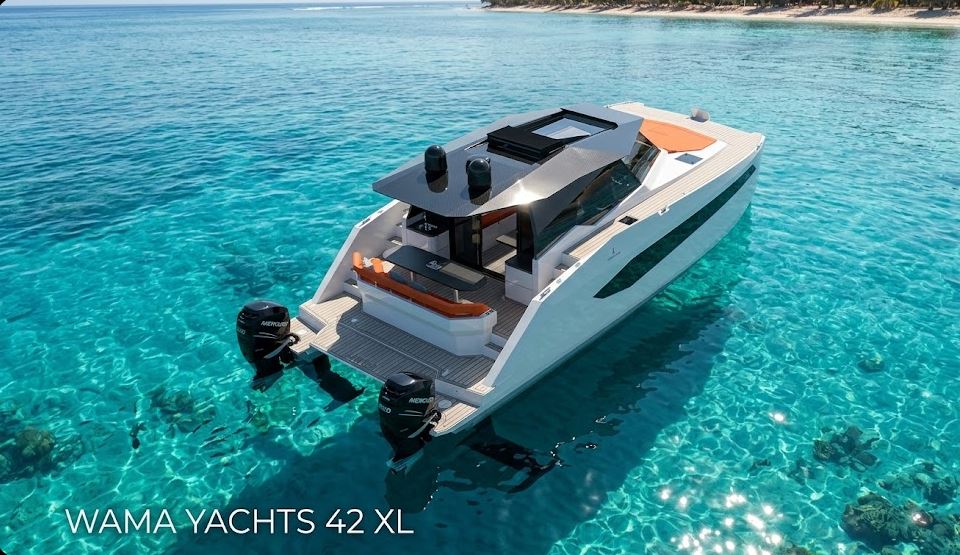 High-angle aerial shot of the Wama Yachts 42 XL luxury power catamaran floating in crystal clear turquoise water. The vessel features a sleek white hull, black superstructure, orange deck cushions, and twin black Mercury outboard engines. A tropical coastline with a white sand beach is visible in the background. Text overlay reads: WAMA YACHTS 42 XL