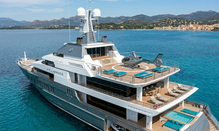 Aerial view of the Stella Maris superyacht in the Mediterranean, showing the top deck with a helicopter landed on the certified helipad, numerous sun loungers with teal cushions, and a stern swimming pool against a mountain backdrop.